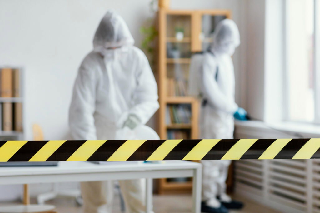 Two people in hazmat suits work behind yellow-black caution tape in a room with shelves and a table, carefully performing trauma restoration.