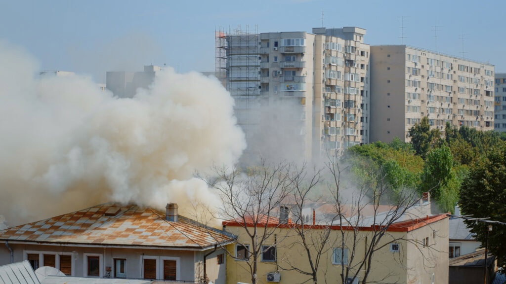 Thick smoke rises from a building fire near apartment blocks in an urban area on a sunny day, highlighting the need for smoke damage restoration and soot removal.